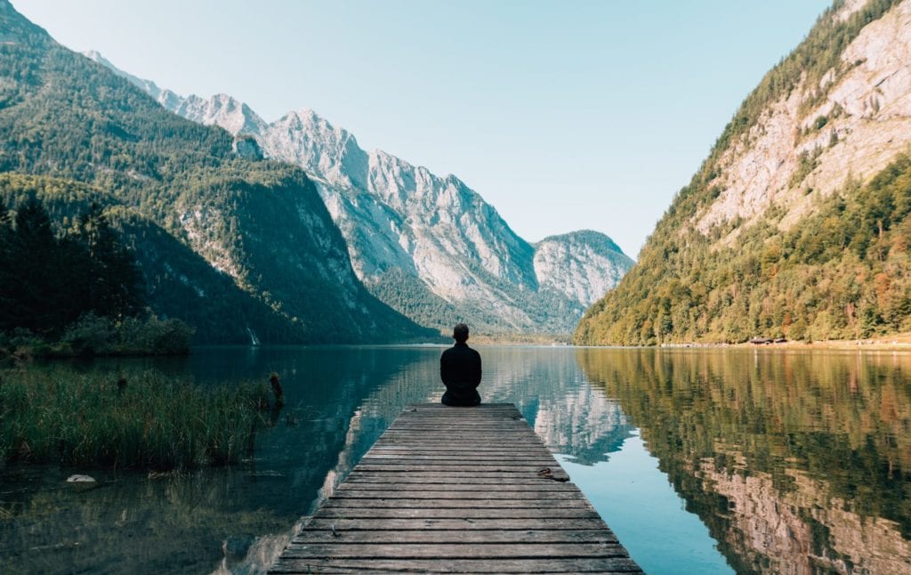 man sitting at the edge of a dock with a beautiful mountain lake background reflected in the water
