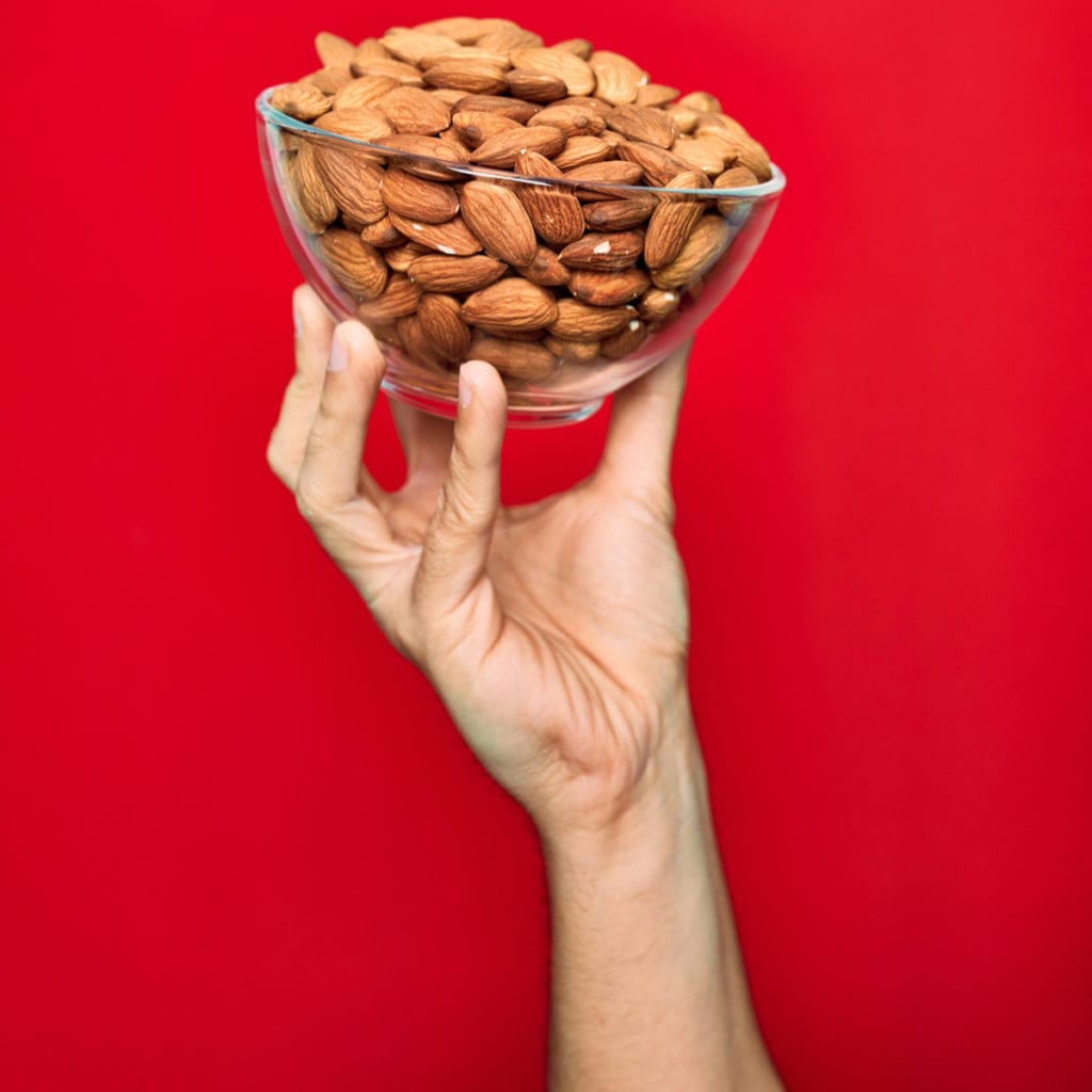 hand holding a bowl of almonds in front of a solid red background