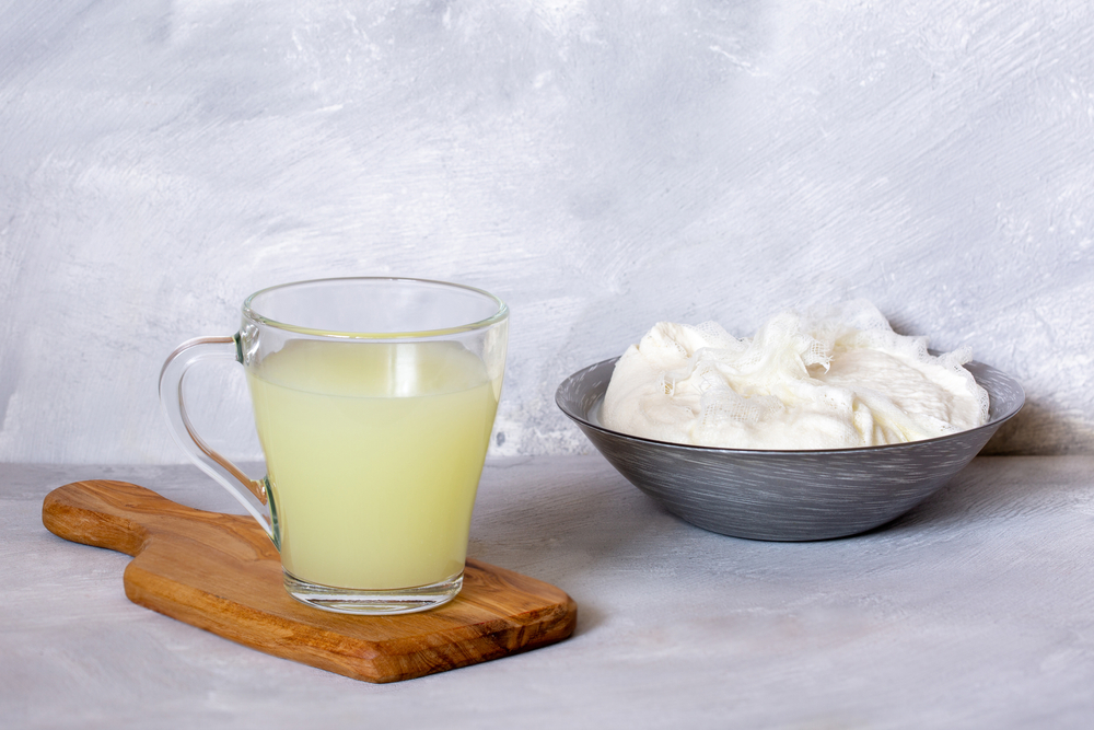 Curds in a bowl, wrapped in cheesecloth, and whey in a clear glass mug sitting on top of a small wooden paddle.