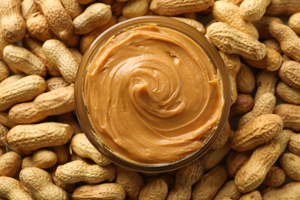An open jar of peanut butter seen from above, surrounded by peanuts in their shells.