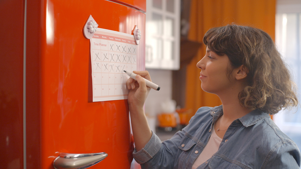 Woman with short, wavy hair and bangs crossing off dates on her calendar hanging on a bright red fridge. The background is blurred, but we can tell it is her kitchen.