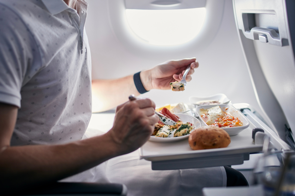 A man sits in a plane, eating food from his pulldown tray stand. He read this Keto FAQ and knows how to stay Keto while traveling!