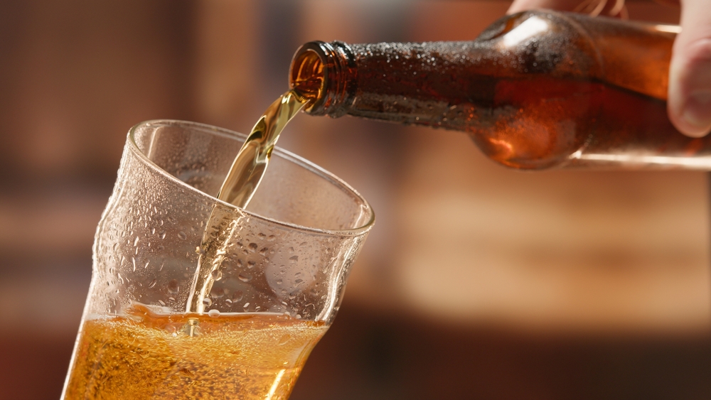 A light beer is poured from a dark brown, perspiring bottle into a wet, water-droplet-lined drinking class. 