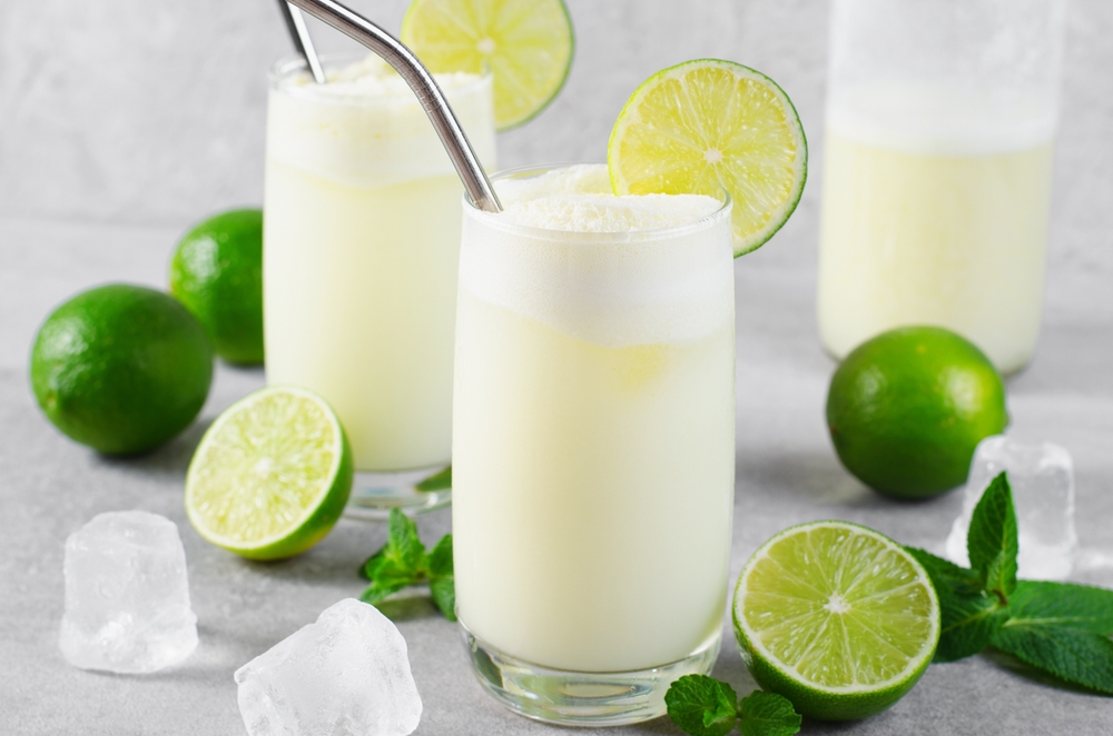 two glasses with Brazilian lemonade topped with and surrounded by limes, with a thick cream on top and two metal straws