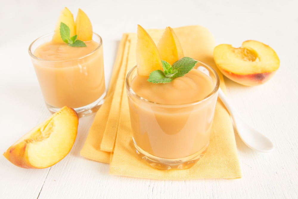 bright picture of two small glasses of a peach smoothie sitting on a yellow napkin with sliced peaches in the background