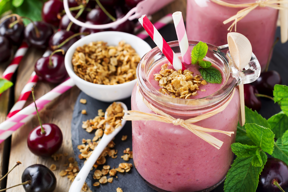 glass of a purple-red smoothie with two straws sticking out and topped with oats and a mint leaf. Background has ripe cherries and a bowl of oats
